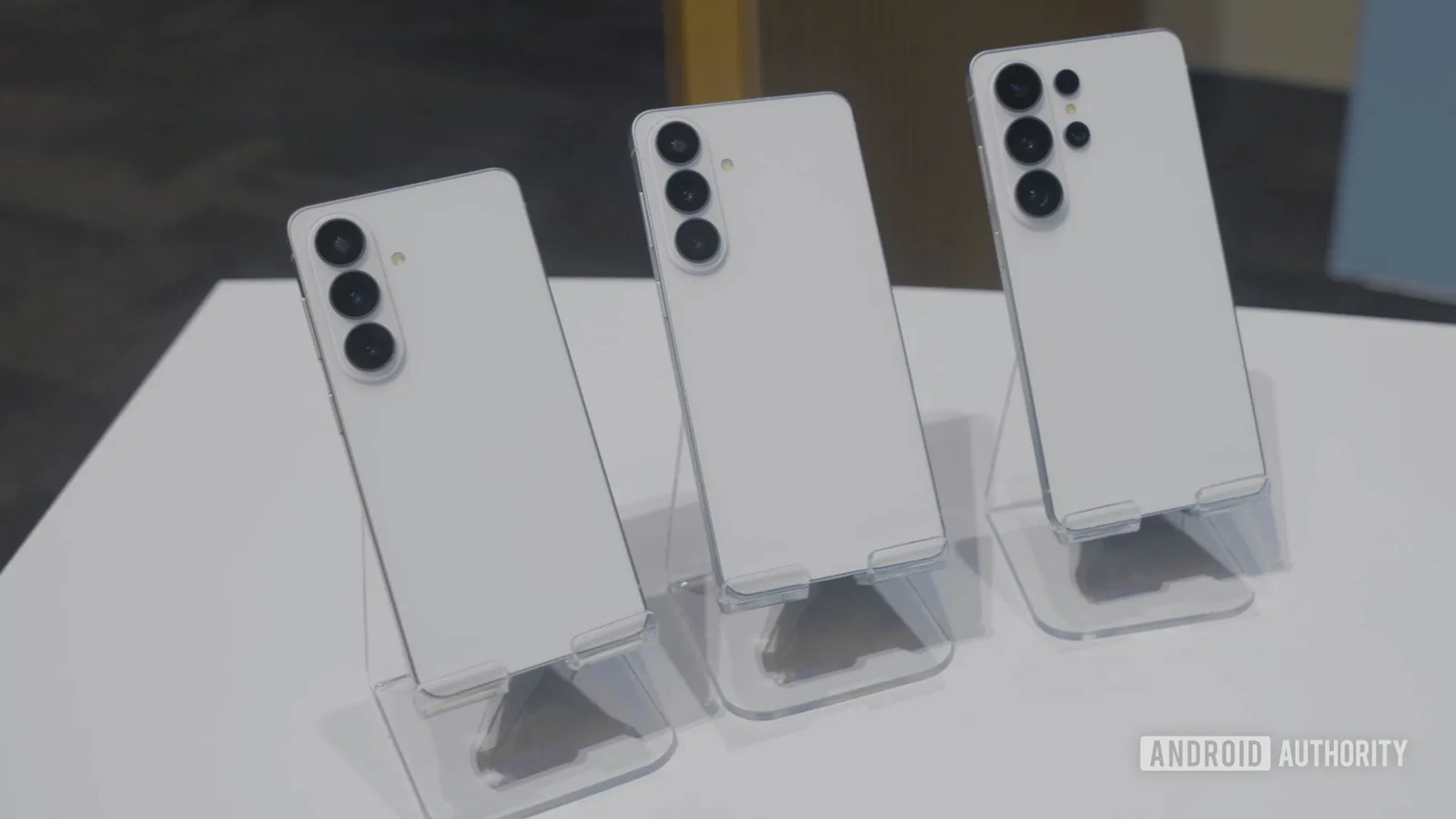 Trei telefoane Galaxy S27 aliniate pe un suport transparent, cu spatele alb.