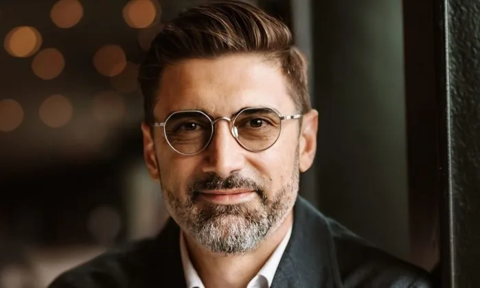 Un bărbat cu ochelari zâmbește, având un look modern și elegant.