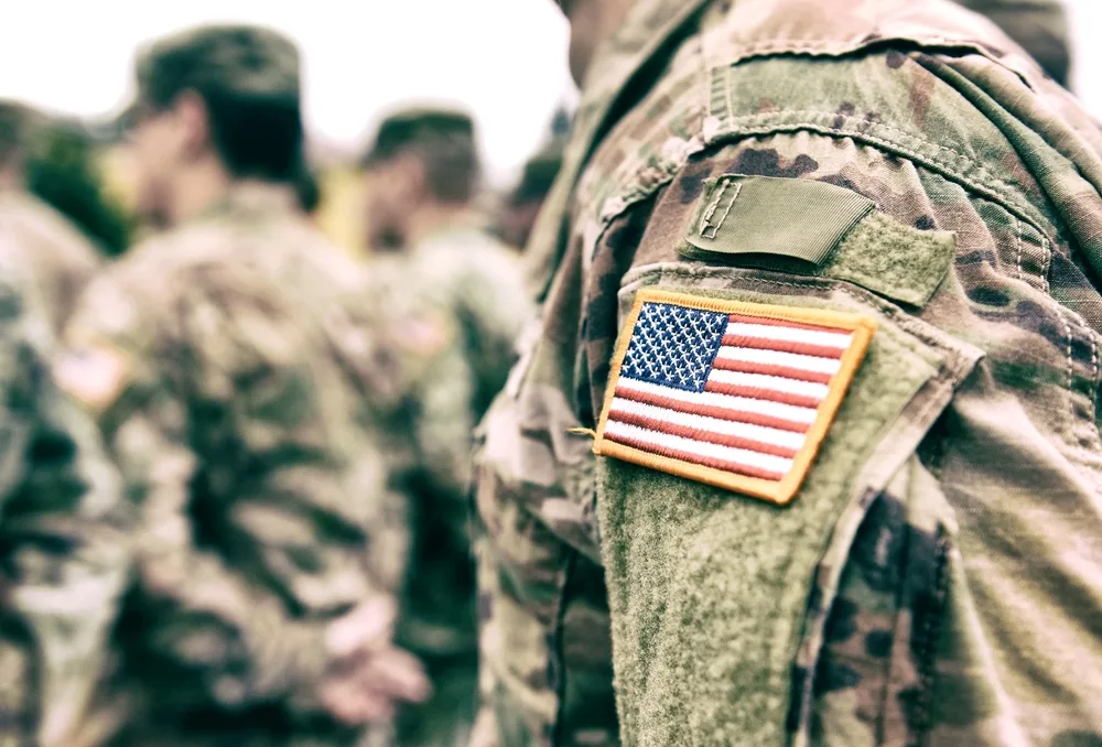 Uniformă militară americană cu steagul SUA pe umăr, soldați în fundal.