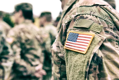 Uniformă militară americană cu steagul SUA pe umăr, soldați în fundal.