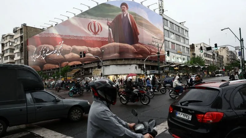 Stradă aglomerată în Iran, cu oameni și vehicule, pe fundal un mural imens.