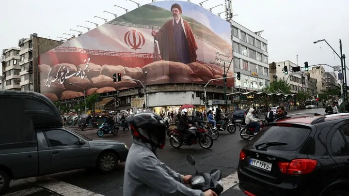 Stradă aglomerată în Iran, cu oameni și vehicule, pe fundal un mural imens.