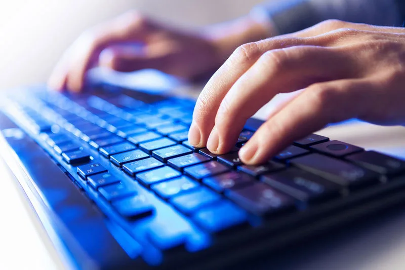 Mâini care tastează pe o tastatură iluminată, simbolizând activitatea în IT.