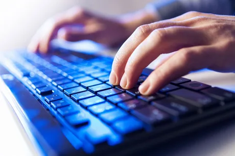 Mâini care tastează pe o tastatură iluminată, simbolizând activitatea în IT.
