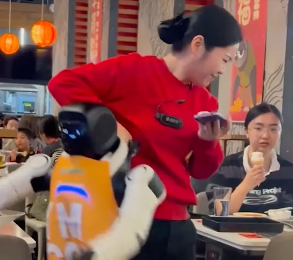 Robot umanoid interacționând cu angajata într-un restaurant aglomerat.