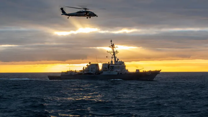 Nava militară însoțită de un elicopter pe fundalul unui apus de soare.