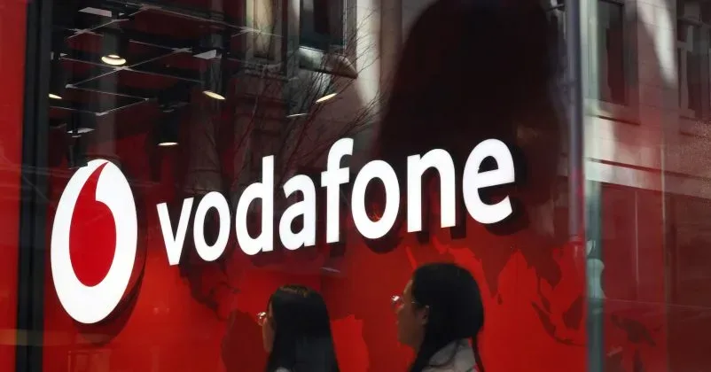 Logo Vodafone pe un fundal roșu, cu oameni trecând pe lângă.
