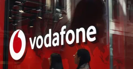 Logo Vodafone pe un fundal roșu, cu oameni trecând pe lângă.