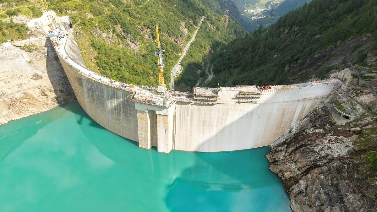 Baraj în construcție, cu apă turcoaz și peisaj montan în fundal.