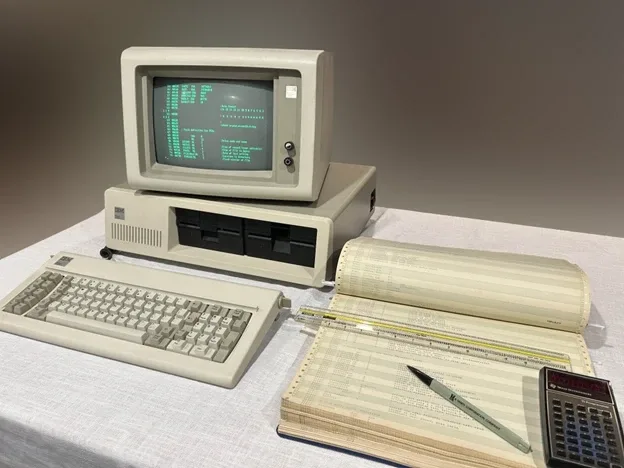 Un computer vechi cu monitor, tastatură și documente tehnice pe masă.