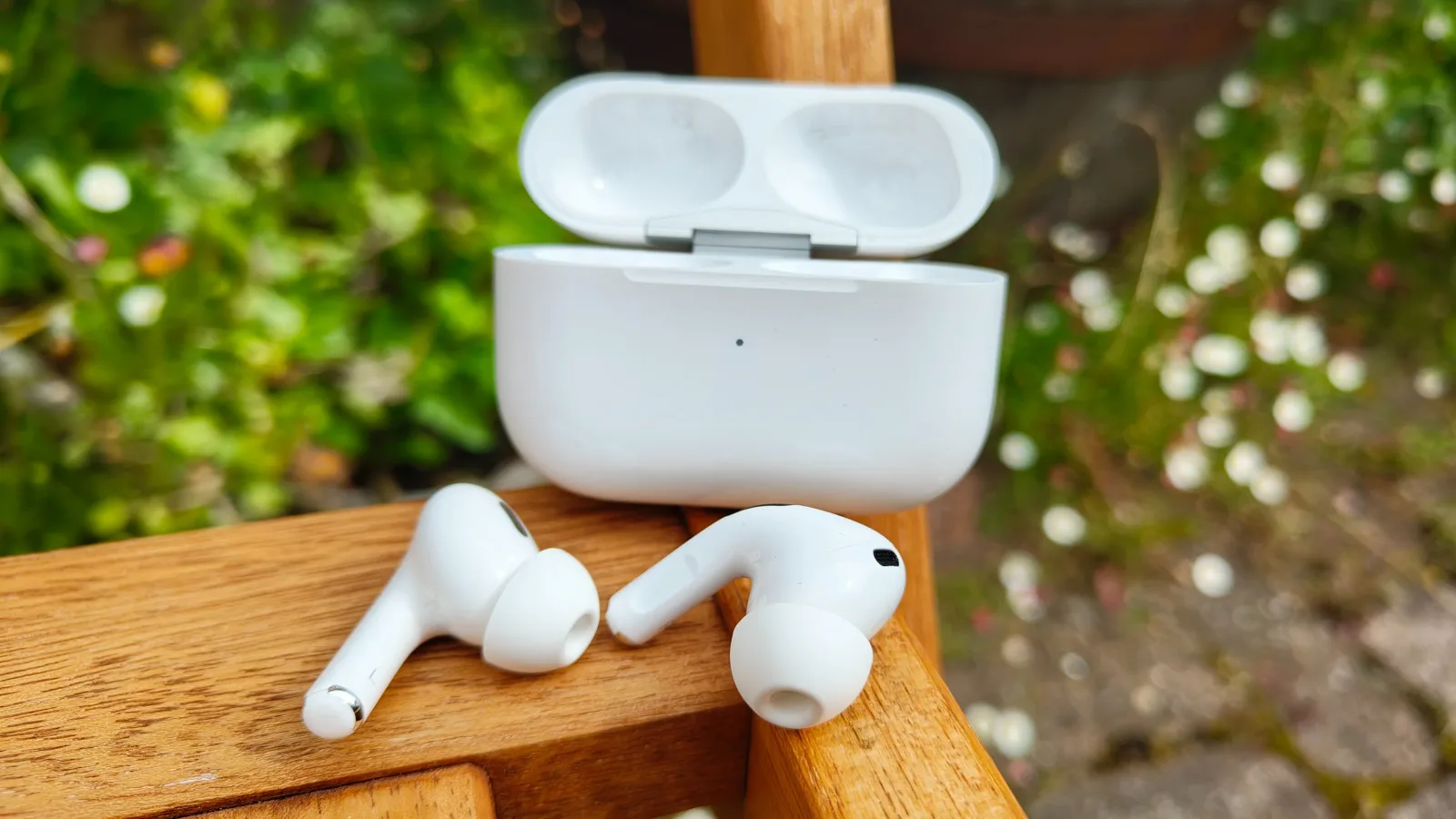 Căști AirPods Pro 3 pe o bancă din lemn, cu flori în fundal.