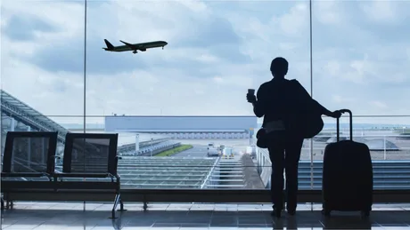 Persoană așteptând un zbor la aeroport, cu un avion în zbor.