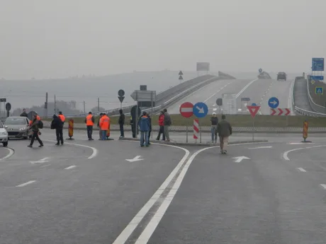 Protest pe șosea, participanți în haine de protecție, vreme mohorâtă.