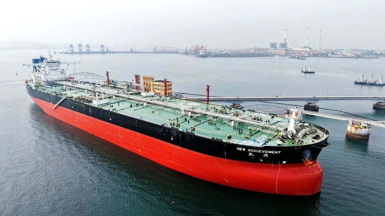 Nava petrolieră navigând pe ape calme, cu porturi în fundal.