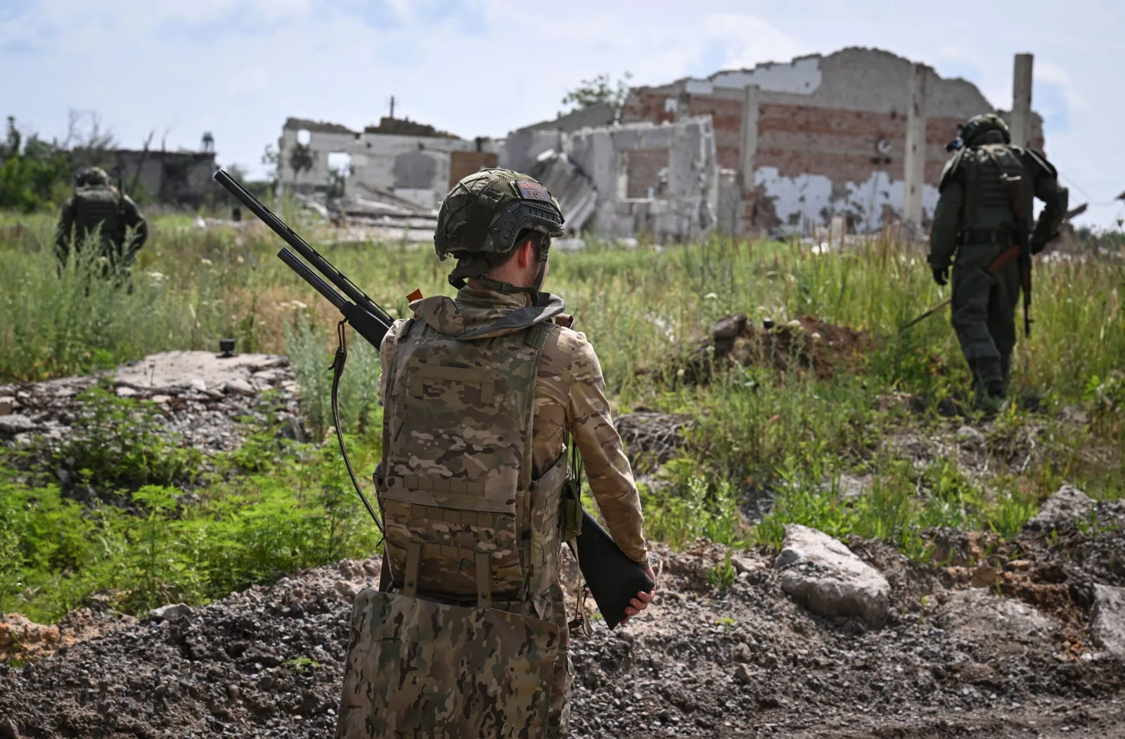 Soldați în terenul afectat de conflict, cu clădiri distruse în fundal.