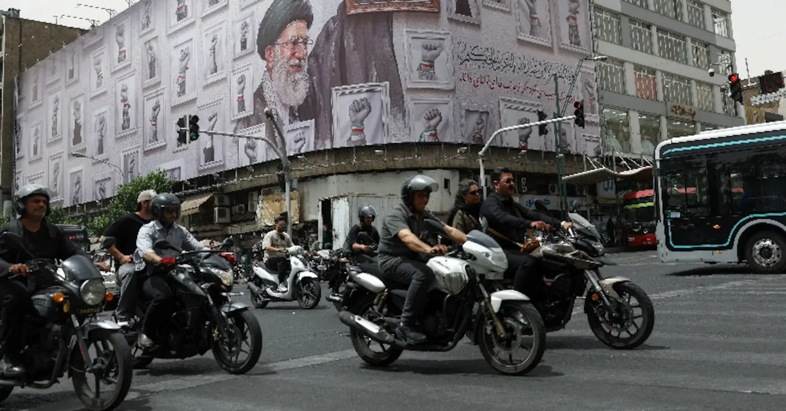Motocicliști circulând pe o stradă aglomerată din Teheran, cu un mural pe fundal.