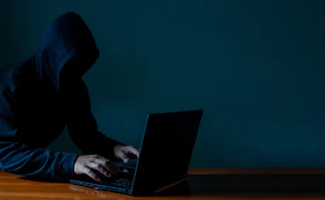 Un individ cu glugă folosește un laptop într-un mediu întunecat.