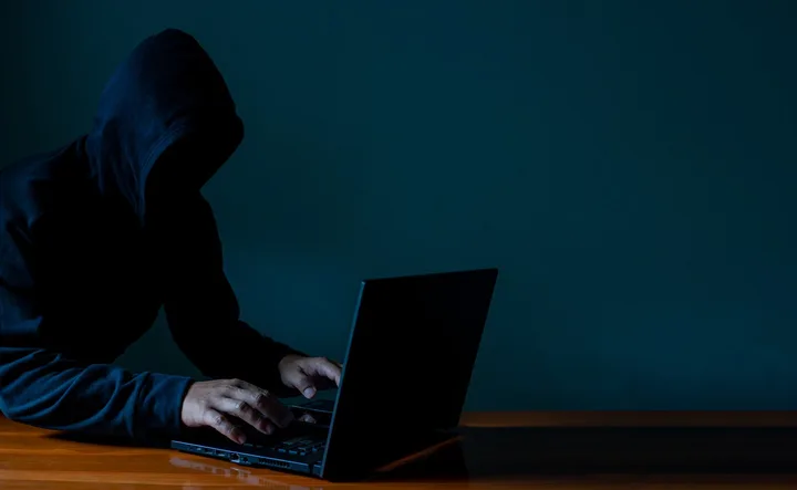 Un individ cu glugă folosește un laptop într-un mediu întunecat.