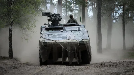 Tanc militar în mișcare printr-o pădure cu praf și vegetație.