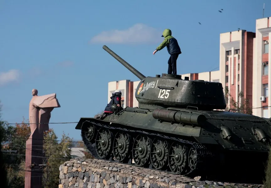 Un copil se joacă pe un tanc, în fundal se vede o statuie și clădiri.