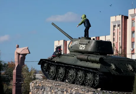 Un copil se joacă pe un tanc, în fundal se vede o statuie și clădiri.