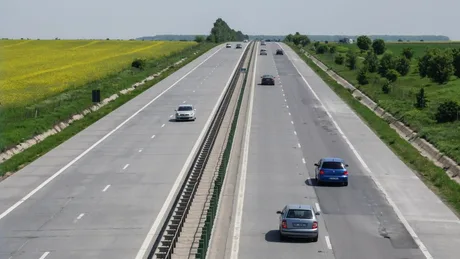 Peisaj de autostradă cu trafic și câmpuri verzi în fundal.