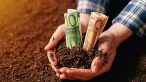 Mână care ține bancnote în pământ, simbolizând sprijinul financiar pentru fermieri.