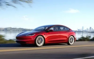 Tesla Model 3 roșie pe un drum lângă apă, cu orașul în fundal.