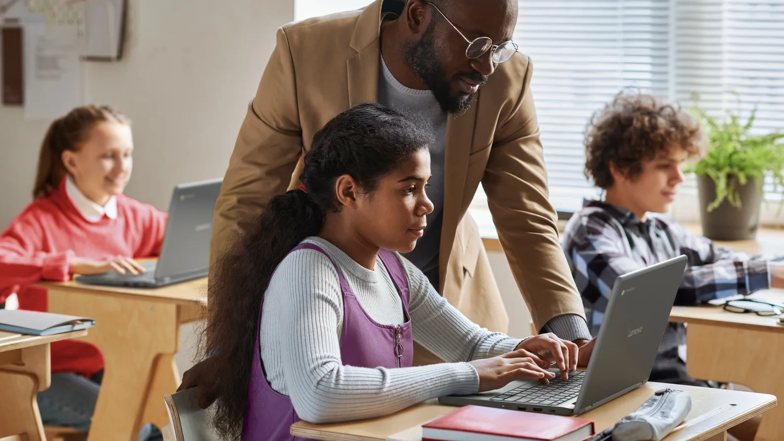 Elevi folosind Chromebook-uri Lenovo în clasă, cu un profesor ajutându-i.