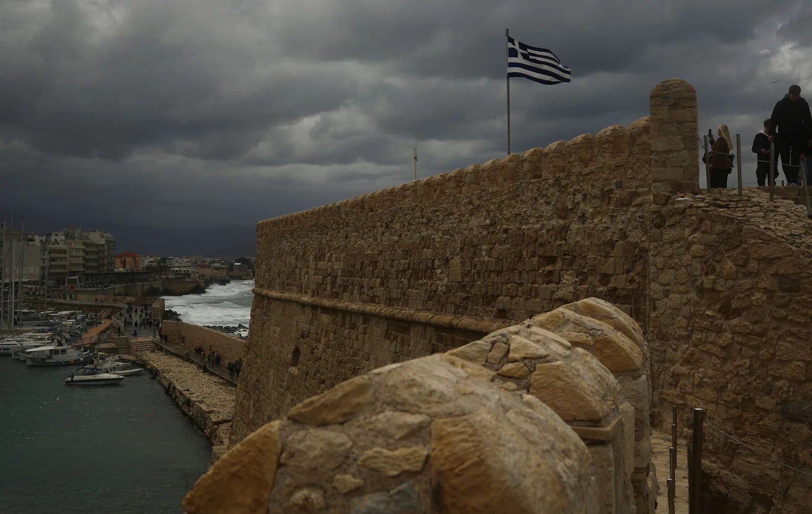 Peisaj de coastă din Creta, cu ziduri de fortificație și steagul Greciei.
