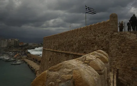 Peisaj de coastă din Creta, cu ziduri de fortificație și steagul Greciei.