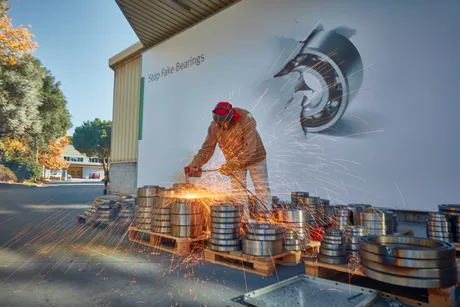 Un muncitor taie metal în fața unei fabrici, cu rulmenți pe fundal.