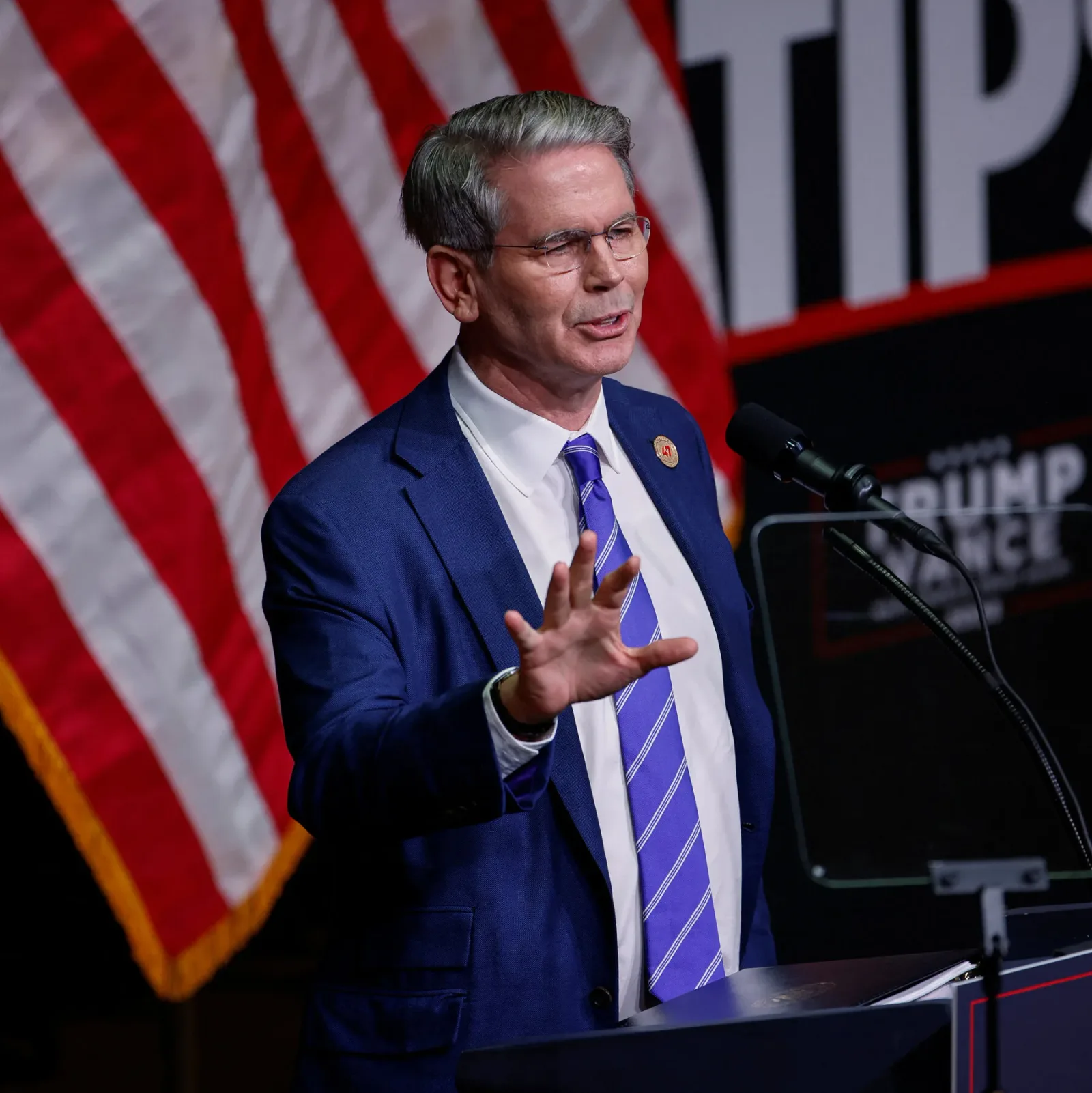 Un politician vorbește la un eveniment public, cu steaguri în fundal.