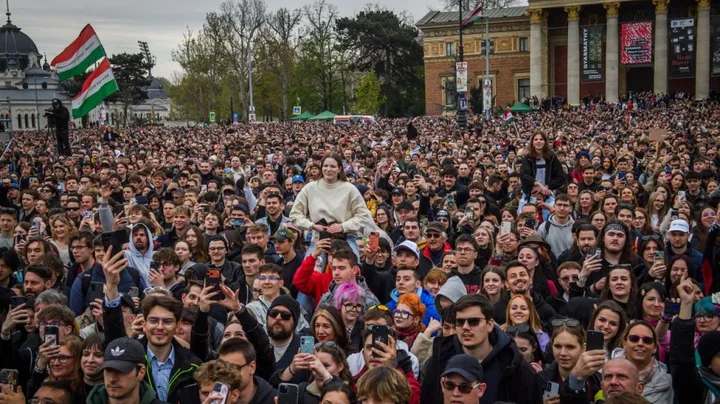 Mulțime de oameni adunați la un eveniment în aer liber, cu steaguri și telefoane ridicate.