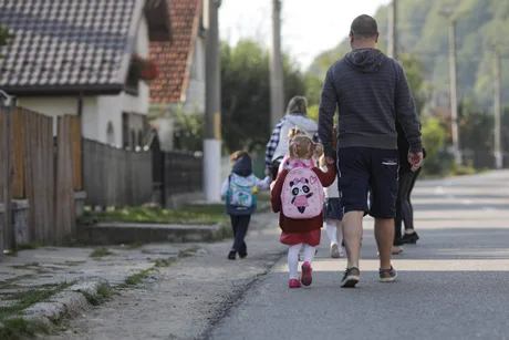 Copii merg spre școală, însoțiți de un adult, cu rucsaci colorati.