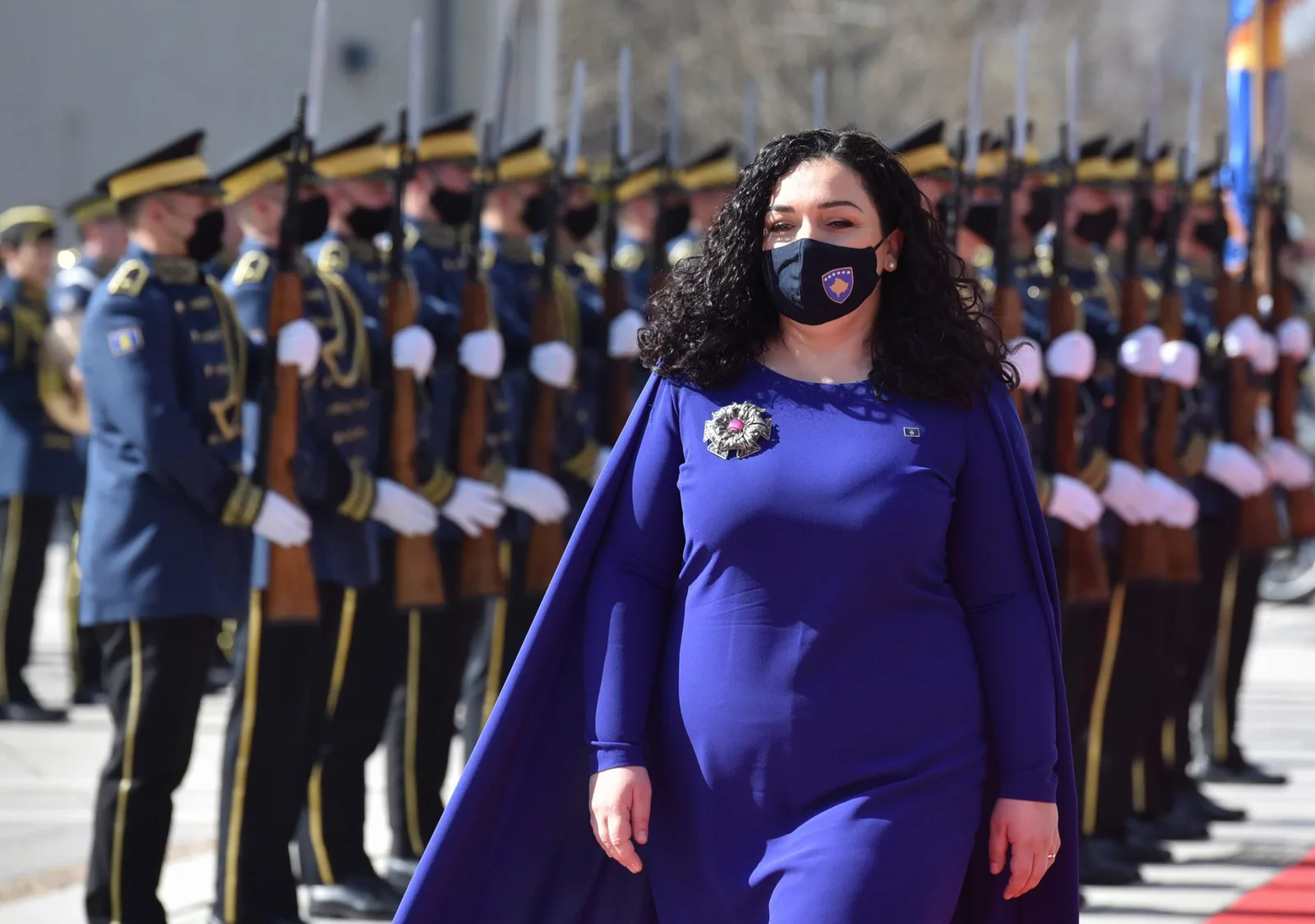 Ceremonie oficială cu soldați în uniformă și o femeie în mantie albastră.