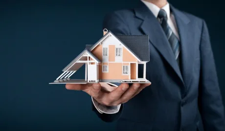 Un bărbat îmbrăcat la costum ține o casă în palmă, simbolizând proprietatea.