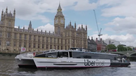 Ambarcațiune Uber Boat navigând pe apă, cu clădiri istorice în fundal.