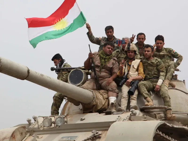 Luptători kurzi pe un tanc, cu steagul Kurdistanului în fundal.