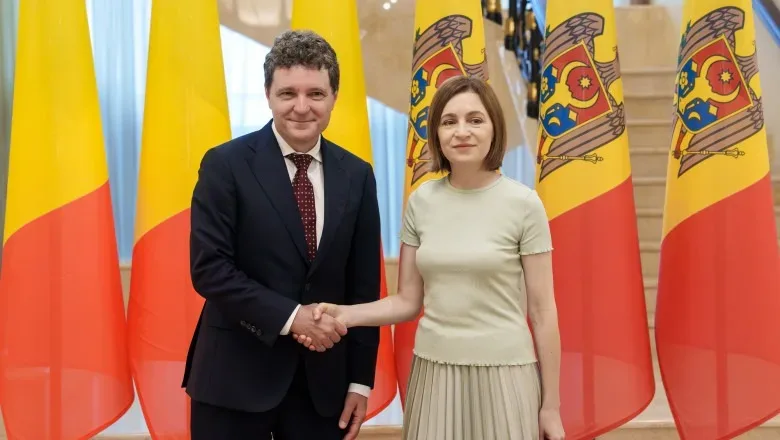 Maia Sandu și un oficial strâng mâna în fața drapelului Moldovei.