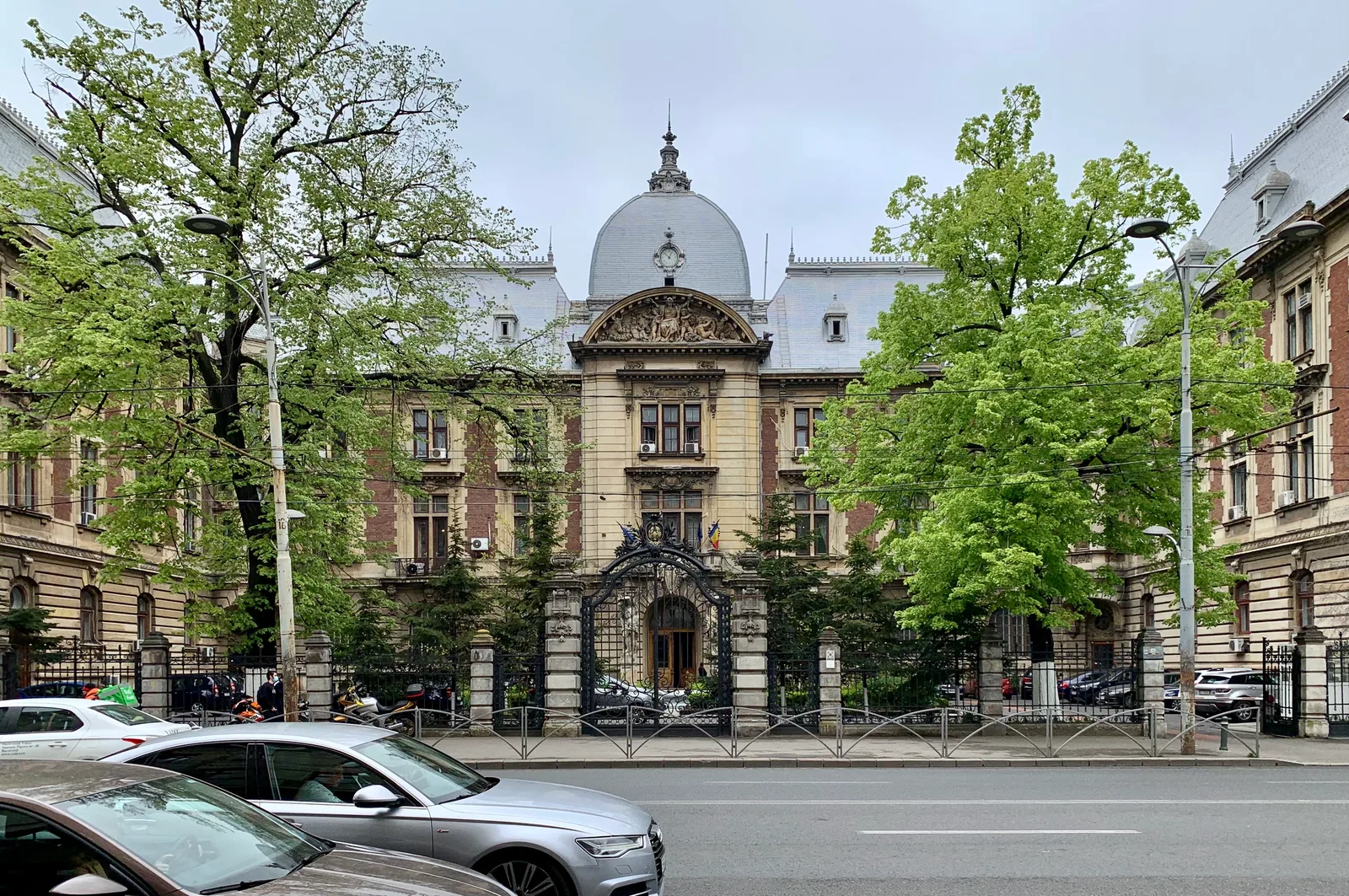 Clădire istorică înconjurată de copaci verzi și trafic urban.