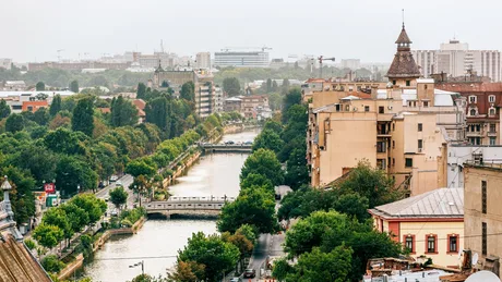 Vedere panoramică a unui oraș cu clădiri și un râu, înconjurat de copaci.