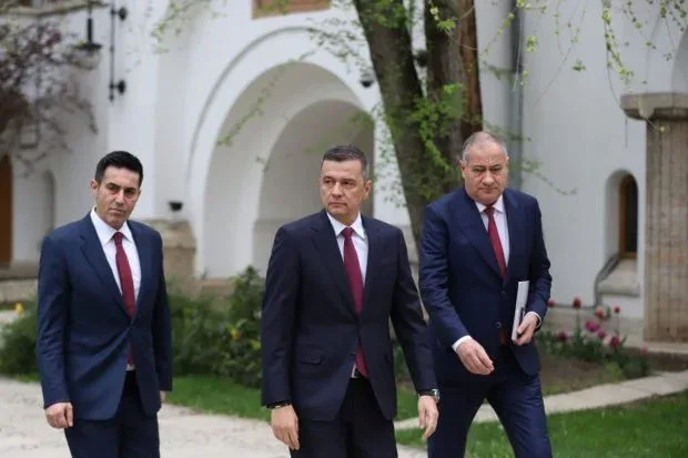 Trei politicieni discută în fața unei clădiri, purtând costume elegante.