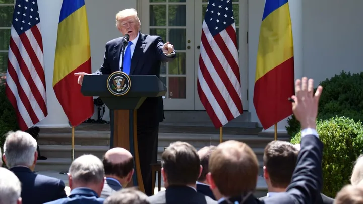Donald Trump susține un discurs în fața jurnaliștilor, cu steagurile României și SUA în fundal.