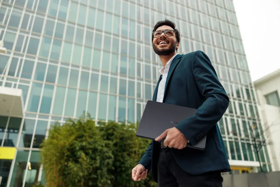 Bărbat zâmbitor cu laptop în mână, în fața unei clădiri moderne.