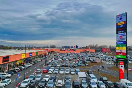 Parcare plină la un parc de retail, cu magazine diverse și cer înnorat.
