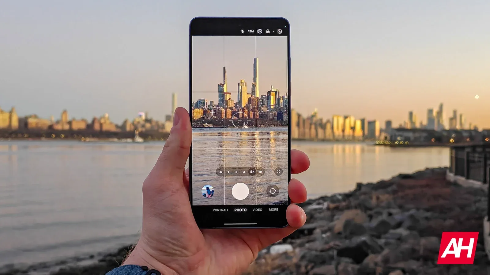 Un smartphone în mână, surprinzând un peisaj urban la apus, cu reflexii în apă.