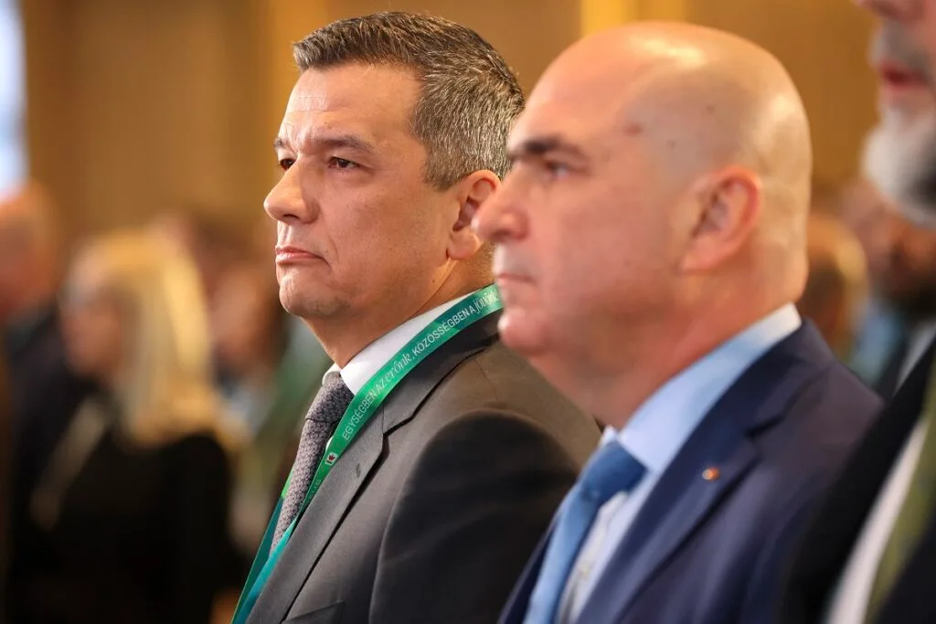 Doi bărbați în costume, ascultând o prezentare la un eveniment oficial.