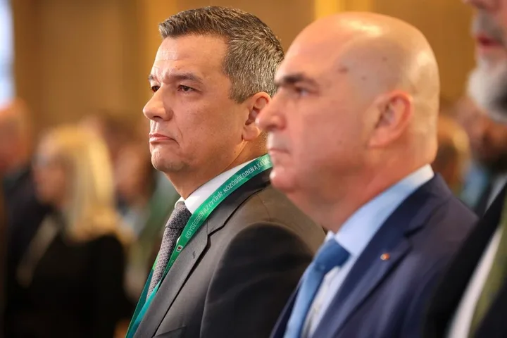 Doi bărbați în costume, ascultând o prezentare la un eveniment oficial.
