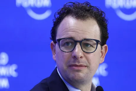 Un bărbat cu ochelari, având un zâmbet rezervat, în fața unui fundal albastru.